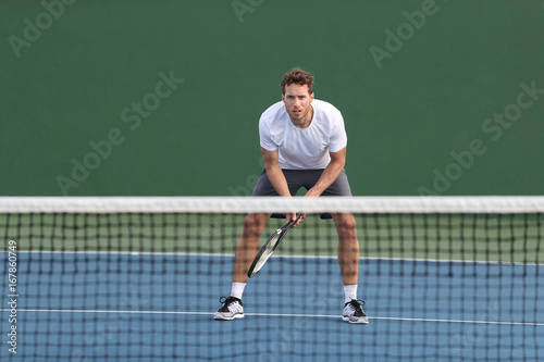 Professional tennis player man athlete waiting to receive ball, playing game on hard court. Fitness person focused behind net ready to return training cardio on outdoor sport activity. © Maridav