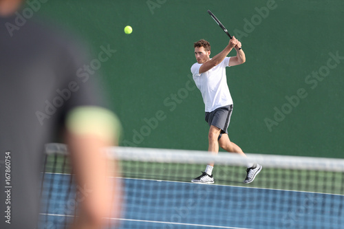 Tennis player man hitting backhand returning ball with racket on green background. Sports men playing together on outdoor court. © Maridav