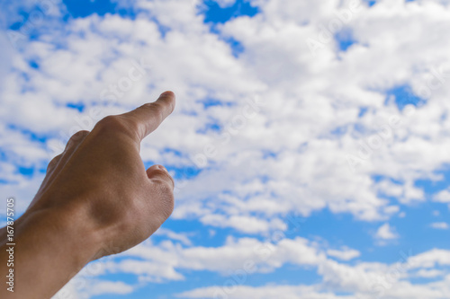 Left forefinger of human hand pointing to blue sky and white clouds.