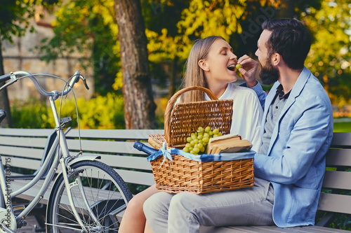 A couple eats grape on a bench in a park.