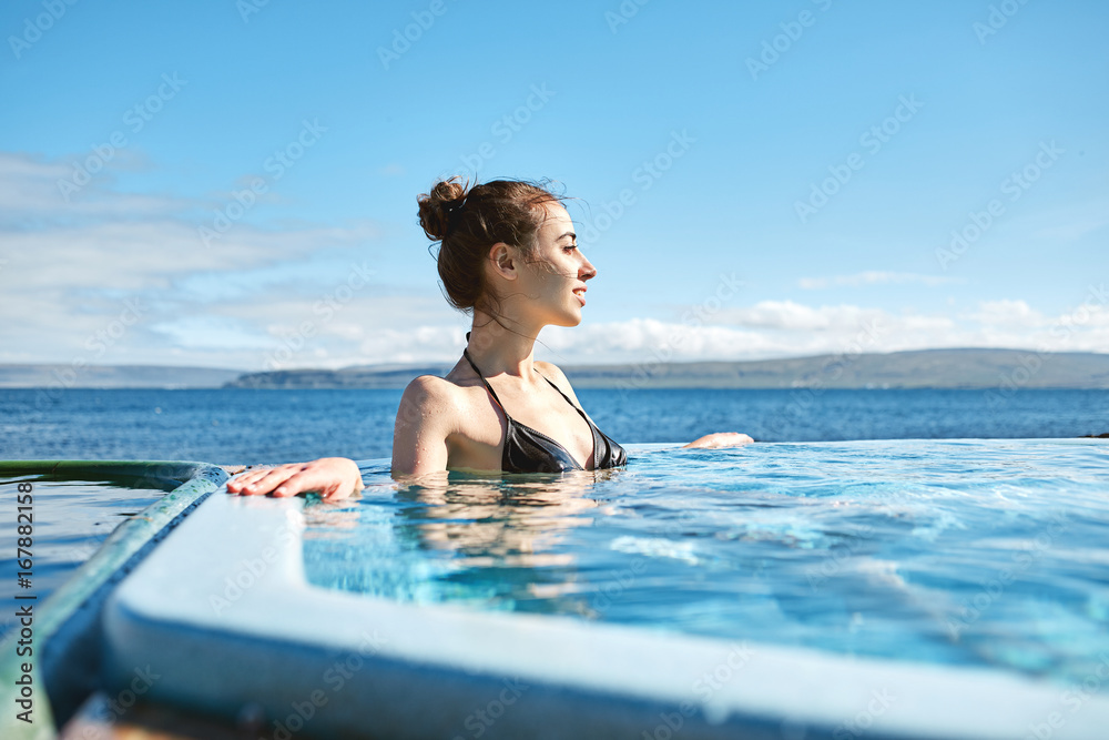 Young cheerful girl swimming in water of pool looking away on background of sea, Iceland, West Fjords.