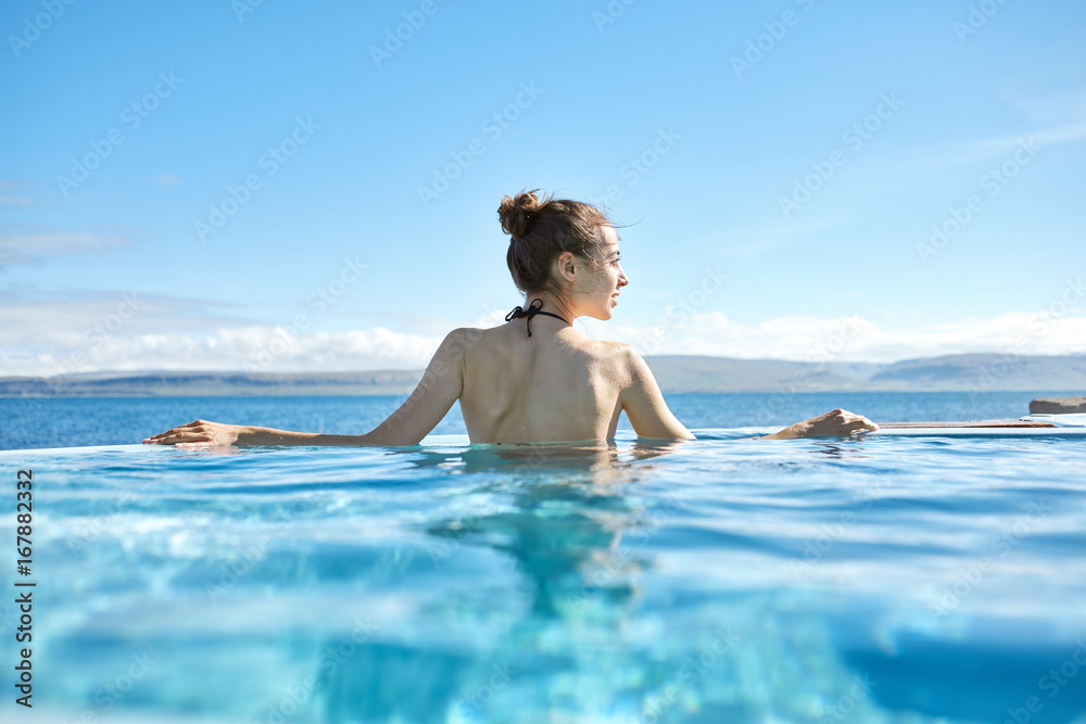 Young cheerful girl swimming in water of pool looking away on background of sea, Iceland, West Fjords. back view