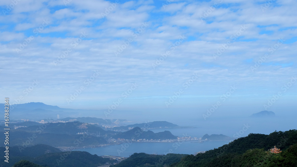 Scenery from the Jilong Mountain, Taiwan, with cloudy blue sky, mountain, sea, and a small temple in the bottom right corner.