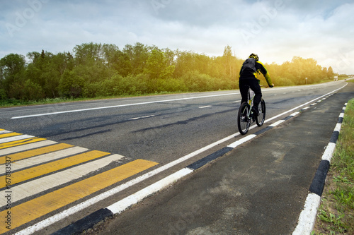 The cyclist is moving along the motorway. A horizontal. A figure of a man on a bicycle moving into the distance in perspective from the left to the right along with yellow and white markings