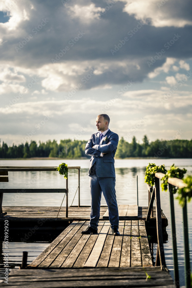 Portrait of young and attractive man in a suit standing on pier next to the huge lake.