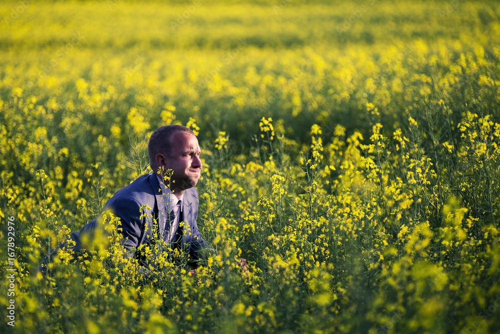 Fototapeta premium Young and attractive man in a suit sitting in yellow rape field and looking somewhere.
