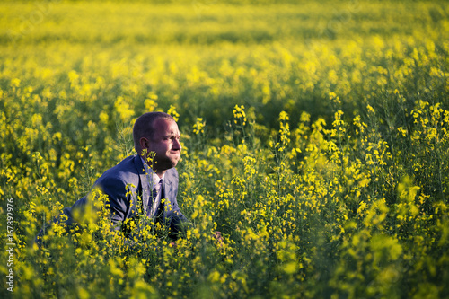 Young and attractive man in a suit sitting in yellow rape field and looking somewhere.