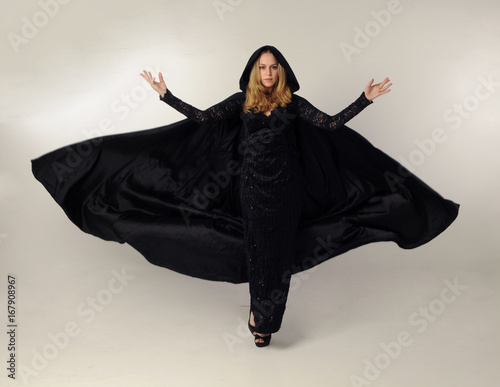 full length portrait of a blonde lady wearing black lace now and hooded cloak, standing pose isolated against creamy background. photo