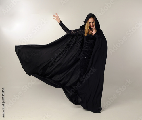 full length portrait of a blonde lady wearing black lace dress and hooded cloak,  standing pose, isolated against creamy studio background. photo