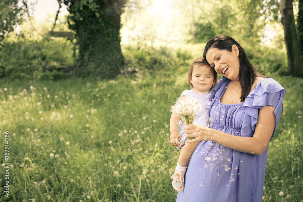 Pregnant mother playing with little daughter in park. Mother and daughter blowing dandelion.