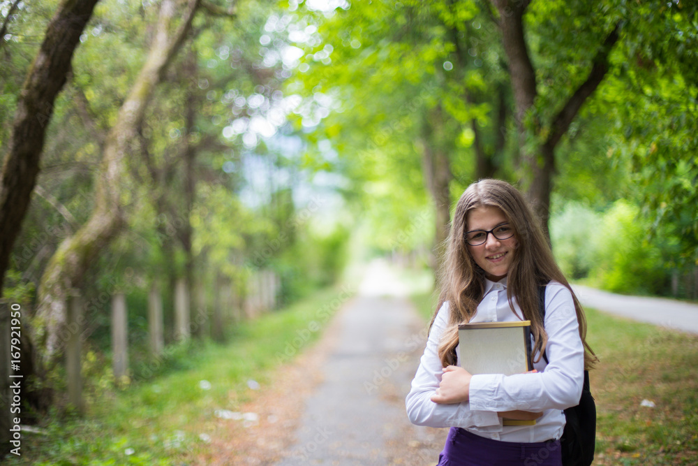 Pretty beautiful blonde child schoolgirl cheerful happy smile holding book back to school enjoying in alley in the nature with backpack, glasses, purple skirt and white shirt Pretty beautiful blonde child schoolgirl cheerful happy smile holding book back to school enjoying in alley in the nature with backpack, glasses, purple skirt and white shirt