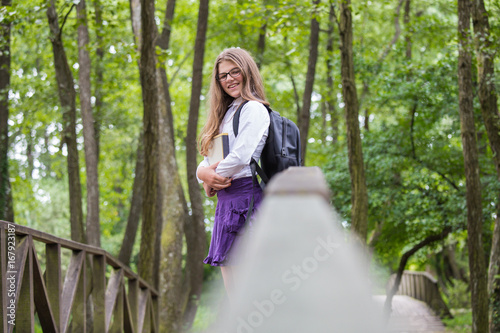 Beautiful pretty blonde school girl child smiling with glasses, white shirt, purple skirt and black backpack leaning on a wooden bridge in nature trees holding books happy back to school autumn