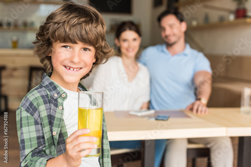 cute curly boy with glass of juice with his parents blurred on background