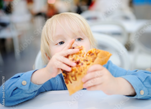 Cute blonde boy eating slice of pizza at fast food restaurant
