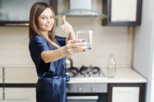 Young happy woman shows "thumbs Up" sign with a glass of drinking water in the kitchen, soft focus