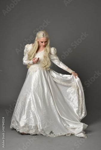 full length portrait of a beautiful blonde woman wearing link white gown, standing pose against a grey background.