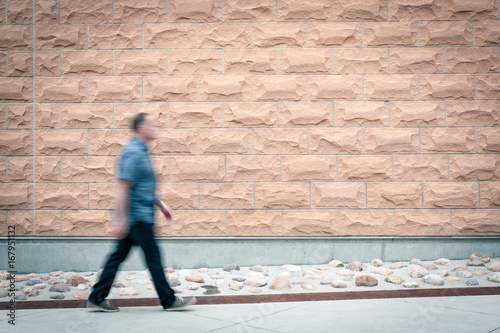 Man walking down sidewalk in front of a large  blank brick rock wall.
