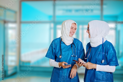 Two female middle eastern doctors in blue medical uniform standing in a hospital hallway, looking at each other, laughing while holding smart phones in their hands