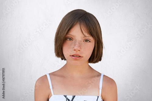 Obraz na plátne Serious freckled girl with bobbed hair and dark eyes looking directly into camera, isolated over white background
