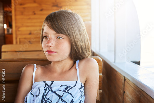 Pensive little child with bobbed hair resting on wooden deck, looking aside while dreaming about something. Pretty girl in white and black dress sitting at wooden bench and table on big ship