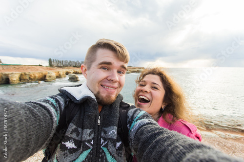 Happy traveling couple in love taking a selfie on phone at the beach on winter or autumn day. Pretty girl and her handsome boyfriend having fun, crazy emotional faces , laughing. © satura_