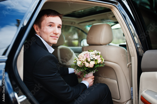 Portrait of a handsome groom sitting in the wedding car with a bouquet in his hands.