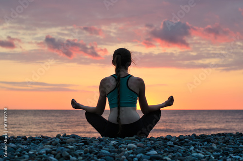 Woman meditating on the beach in lotus position. View from the back, sunset