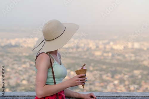 Girl in a hat with ice cream in hands looks at the city from a height photo