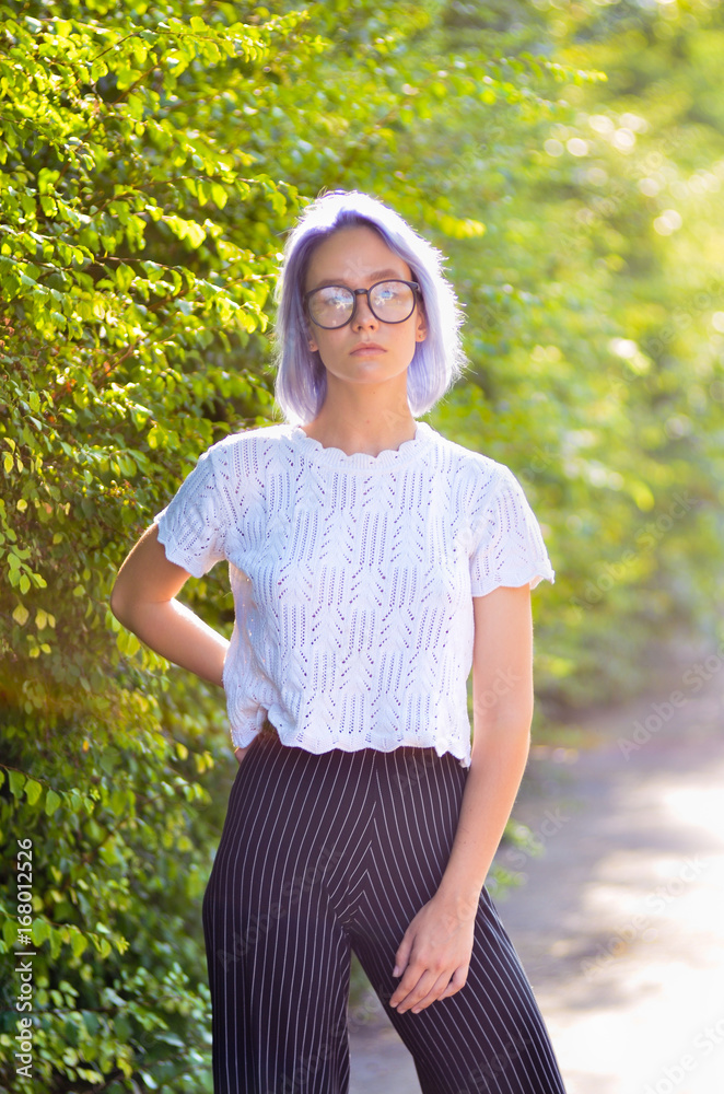 Αυτοκόλλητο Young informal hipster girl with ashen purple hair in glasses posing in front of a camera in the park on a background of green bushes