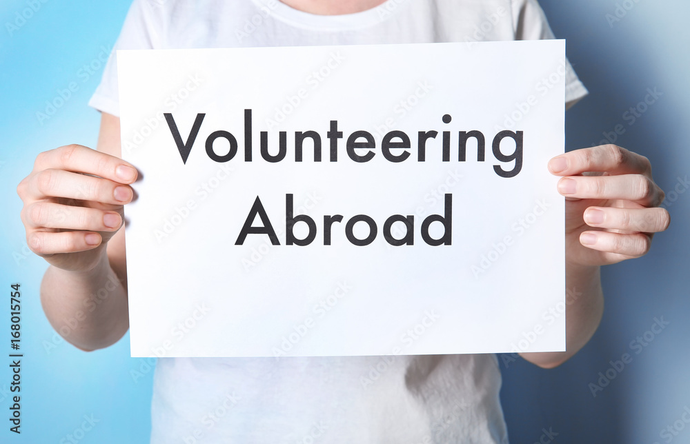 Φωτογραφία Woman holding sheet of paper with text VOLUNTEERING ABROAD on color background
