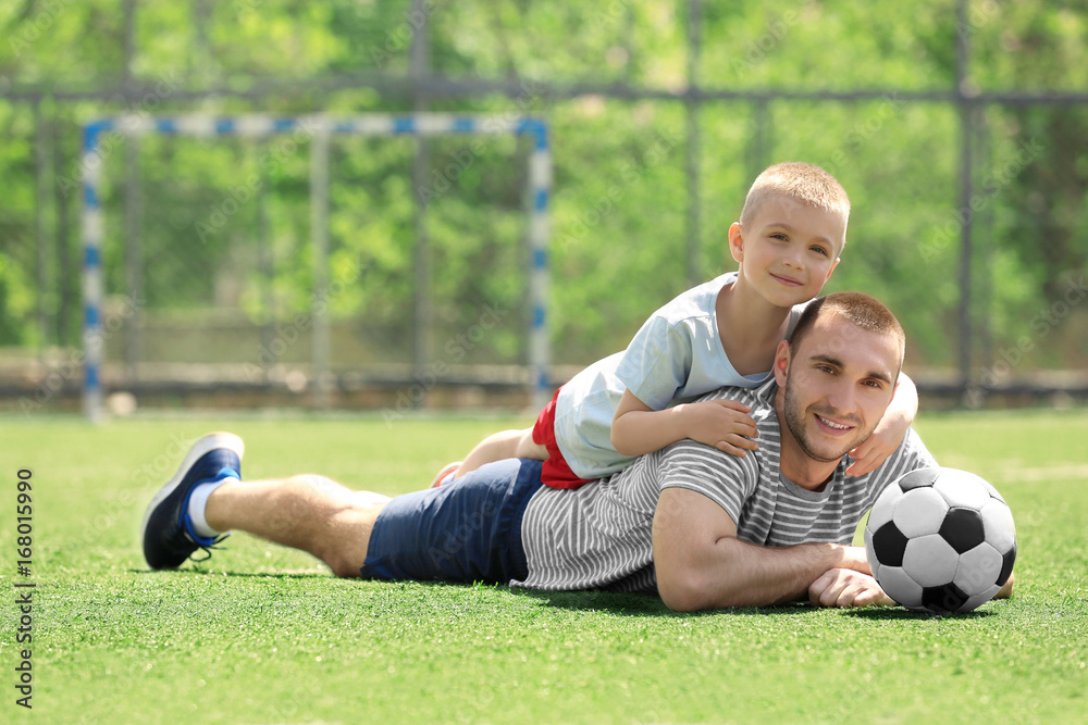 Father and son with ball lying on soccer pitch