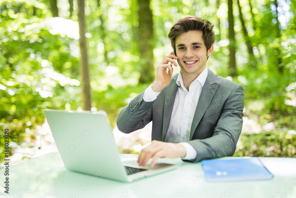 Portrait of young handsome business man sitting at office desk in the green park or forest and work at laptop while talk at phone with costumer. Business concept. Portrait of young handsome business man sitting at office desk in the green park or forest and work at laptop while talk at phone with costumer. Business concept.