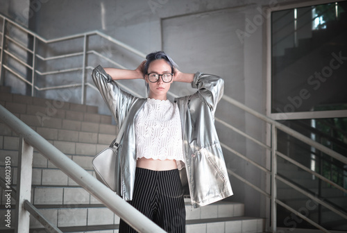 Extraordinary hipster girl with ashen blue hair is dressed in a cloak with a silvery fabric and a backpack on her shoulders posing on the street wearing glasses in the background of the steps. photo