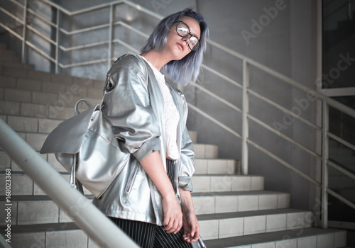 Konstfotografi Space style girl with ashen blue hair is dressed in a cloak with a silvery fabric and a backpack on her shoulders posing on the street wearing glasses in the background of the steps of giving