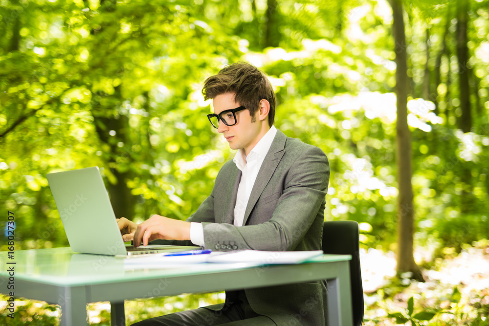 Side portrait of young handsome business man working at laptop at office table in green park. Business concept. Side portrait of young handsome business man working at laptop at office table in green park. Business concept.