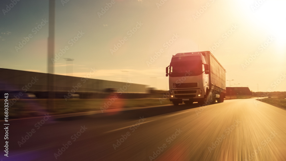 White Semi Truck with Cargo Trailer Attached Drives on the Empty Road. Industrial Warehouses by the Sides of the Highway. Sunset. Blur motion.
