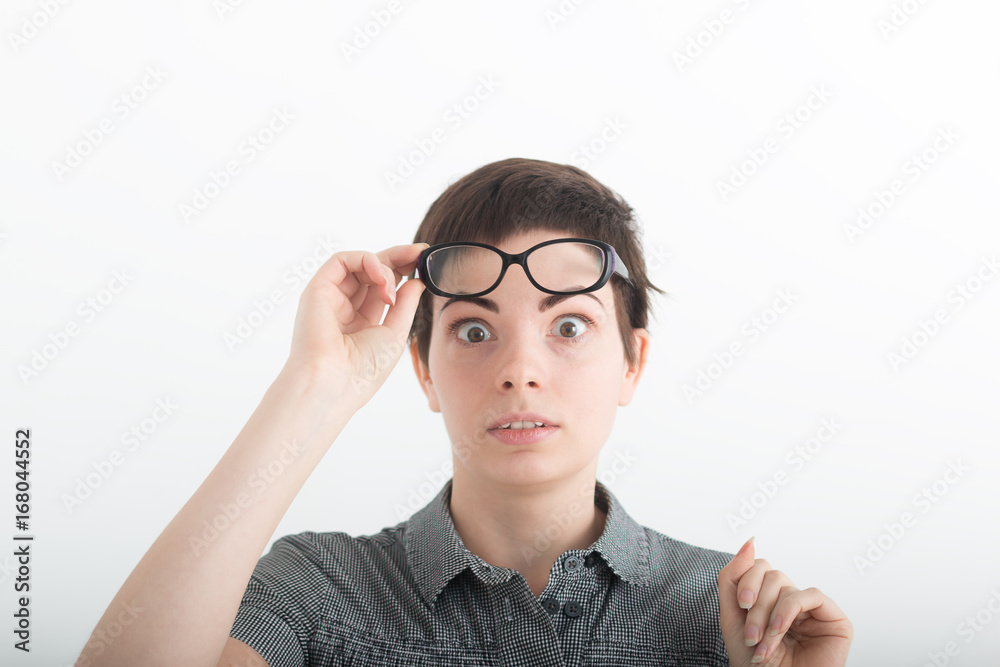 Young beautiful dark-haired woman in grey shirt with astonishment looks upwards, against white studio background. Breaking news. Surprised pleasant woman holding his glasses on forehead