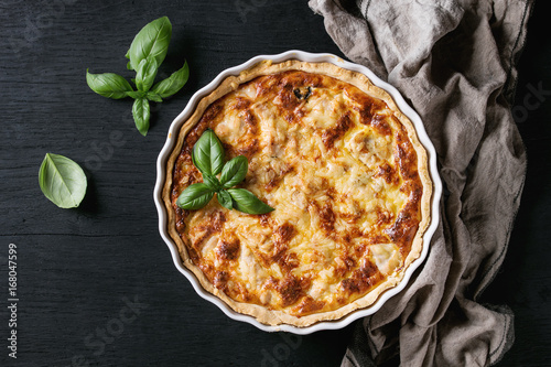 Baked homemade quiche pie in white ceramic form served with fresh greens and textile towel on black wooden burned background. Flat lay with copy space photo