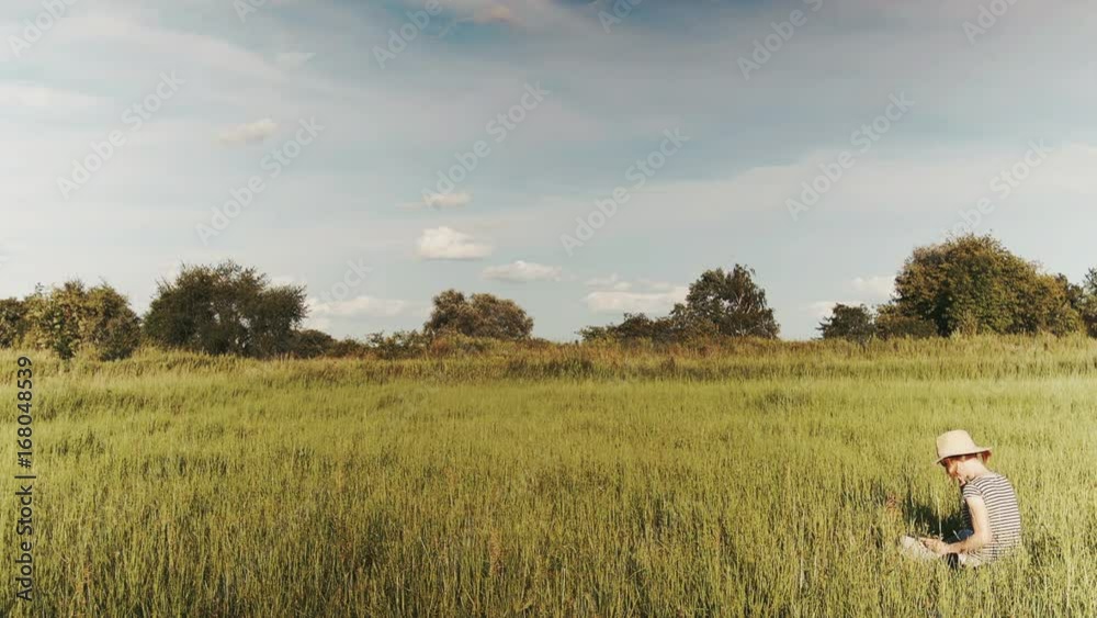 Woman with sunhat sitting in the field with her tablet - wide establishing shot Woman with sunhat sitting in the field with her tablet - wide establishing shot