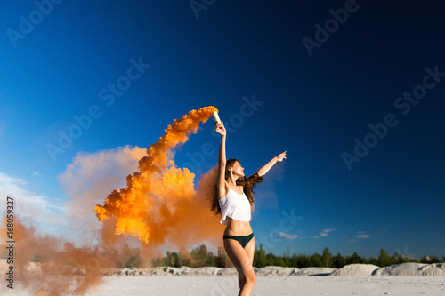 Woman walks with orange smoke on white beach under blue sky