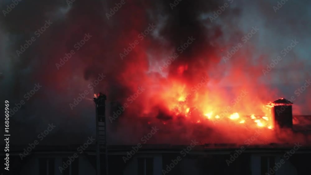 Burning fire flame with smoke on the apartment house roof in the city, firefighter or fireman on the ladder extinguishes fire