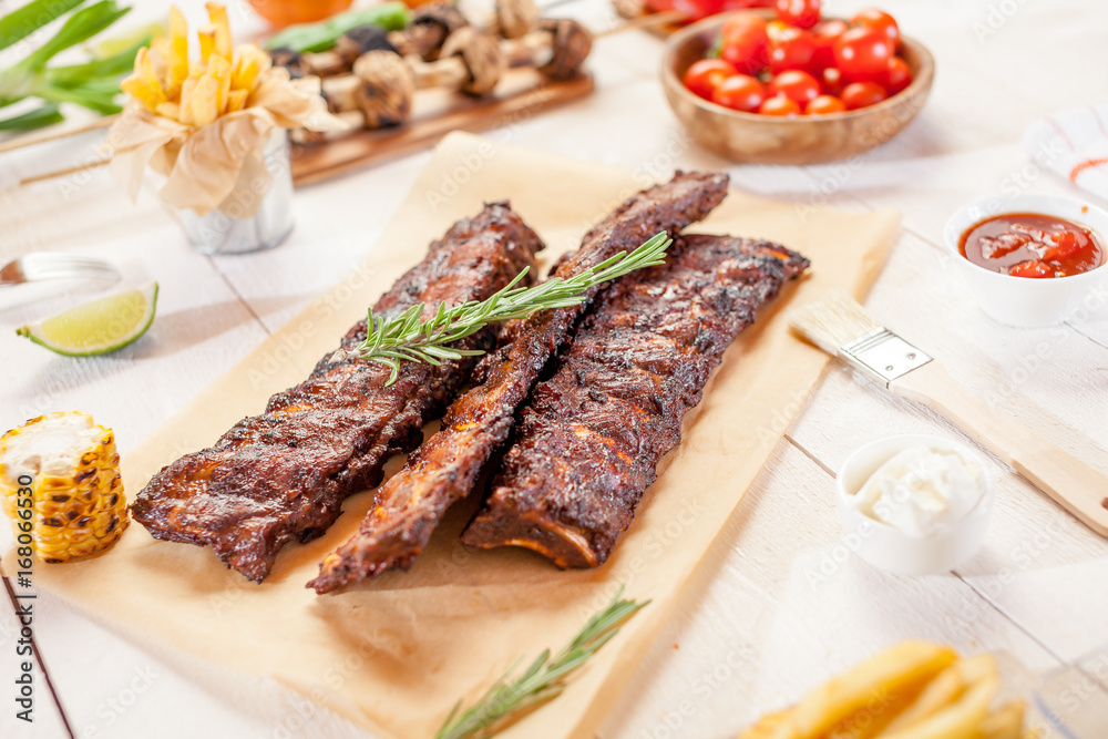 Spicy hot grilled spare ribs from a summer BBQ served with chips, corn and fresh tomatoes on an old wooden cutting board