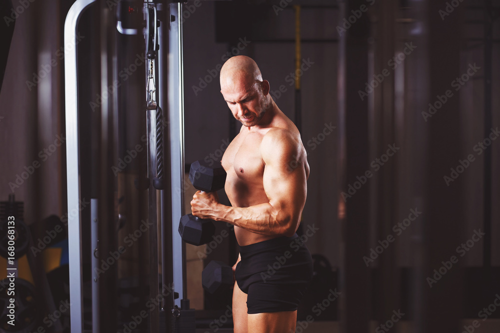 Strong ripped bald man with huge muscles pumping iron. Sports man working out with dumbbells in gym, toned photo. Sport, fitness, bodybuilding concept Strong ripped bald man with huge muscles pumping iron. Sports man working out with dumbbells in gym, toned photo. Sport, fitness, bodybuilding concept