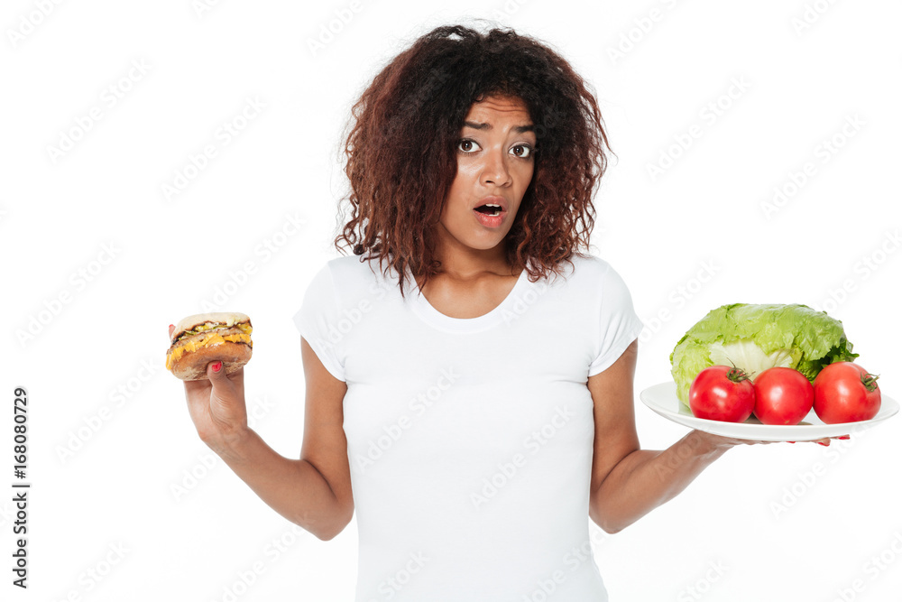 Confused young african woman choosing between burger and vegetables. Confused young african woman choosing between burger and vegetables.