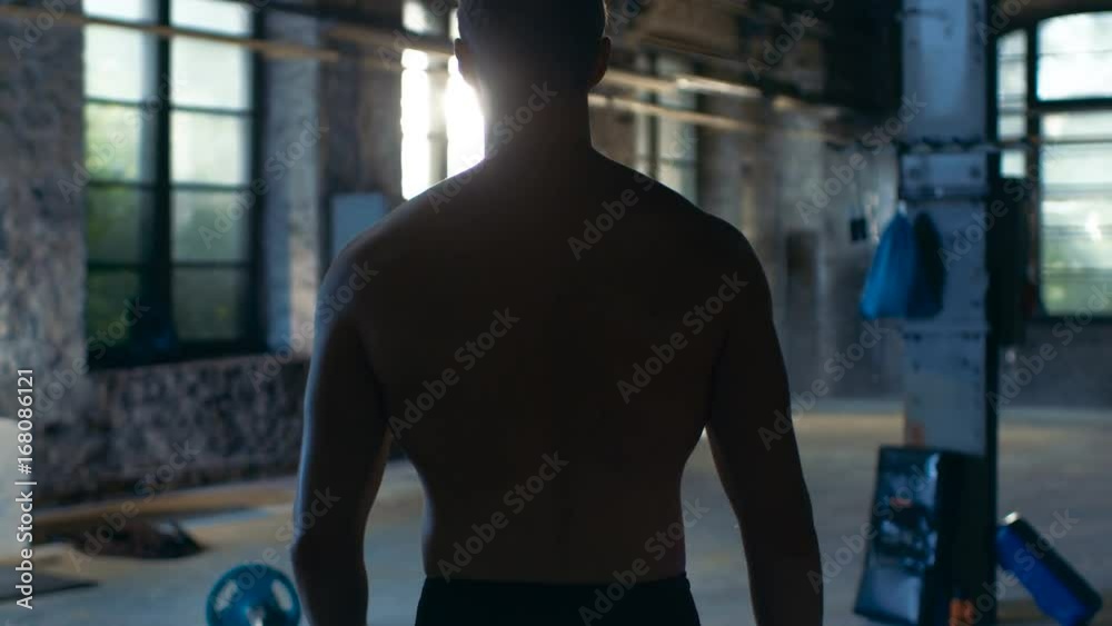 Follow-up Shot of Muscular Shirtless Man Entering Gym in Slow Motion. He's Confident, Building is Industrial and Hardcore, Various Cross fitness/ Bodybuilding Equipment lying on the Floor.