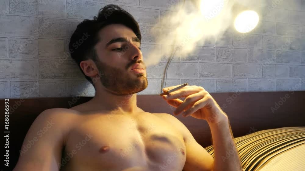 Topless good-looking fit young man smoking cigarette in bed at home before going to sleep. Dangerous situation. Slow motion.
