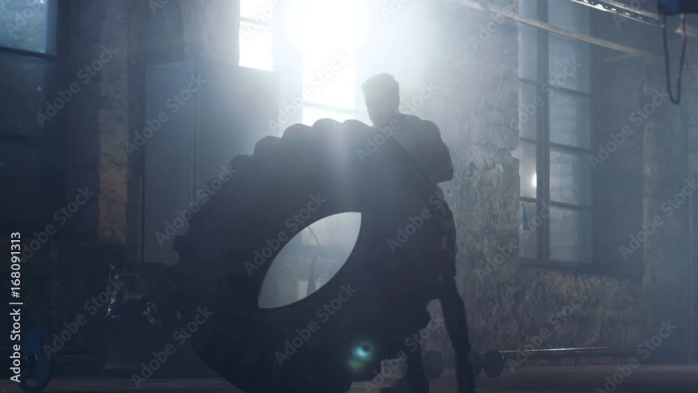 Strong Muscular Man Lifts Tire as Part of His Bodybuilding/ Cross Fitness Training. He's Covered in Sweat and Works out in a Abandoned Factory Remodeled into Gym. 