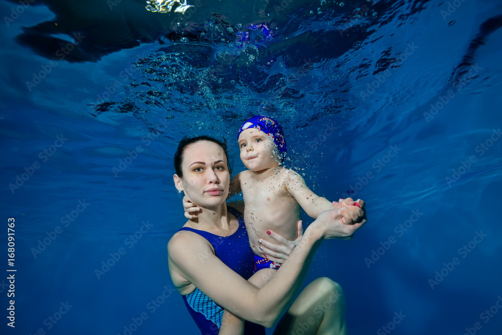 Mother is a swim instructor with my son underwater in the pool on a blue background posing and looking at the camera. Portrait. The view from under the water. Landscape orientation