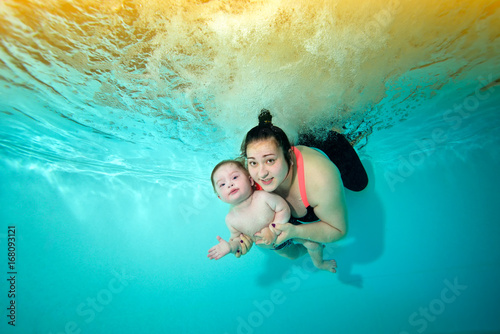 Little boy and mother hug and swim together underwater against the bright lights and looking at me. Portrait. The view from under the water. Landscape orientation