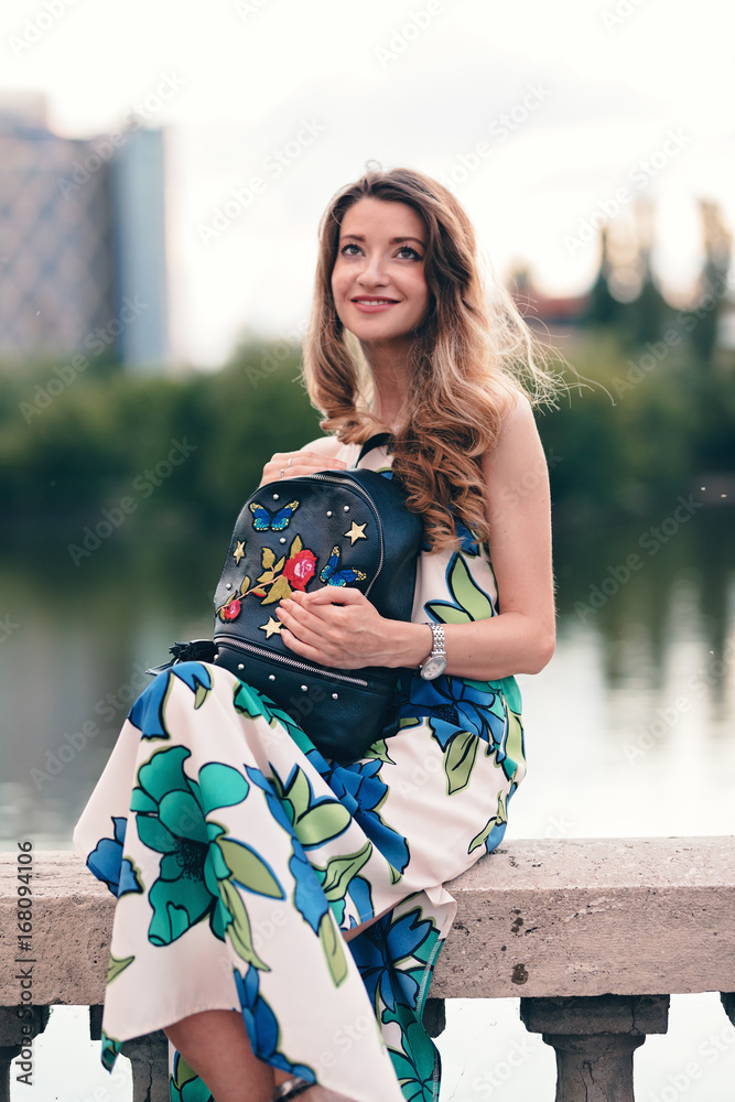 Young teen girl sitting on a concrete handrail. Wearing maxi dress outfit  having floral patterns. Looking at the evening pleasant sky. Holding a  designed black backpack. Lake in background. Stock Photo